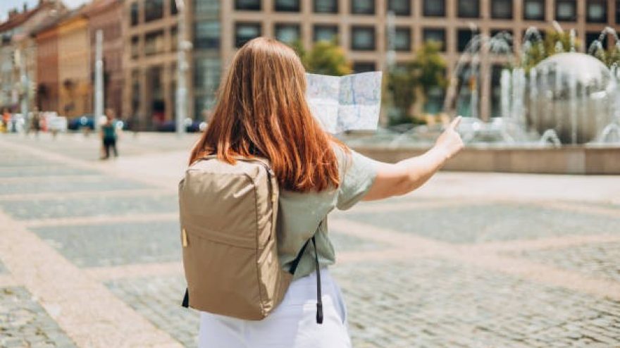 Attractive young female tourist is exploring new city. Redhead woman pointing finger and holding a paper map in a warm sunny day. Urban lifestyle concept.