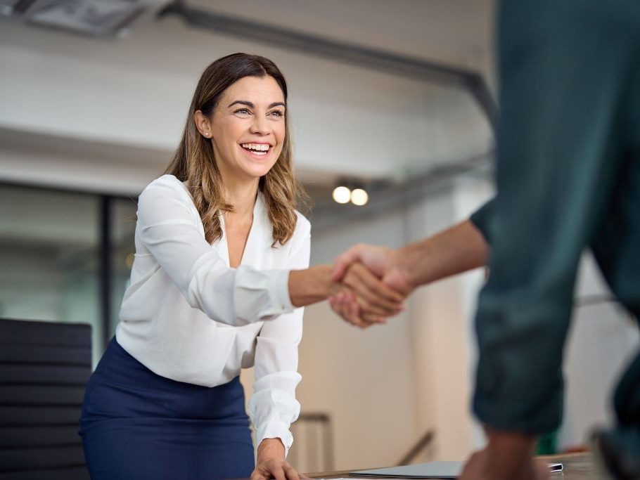 Happy mid aged business woman manager handshaking greeting client in office. Smiling female executive making successful deal with partner shaking hand at work standing at meeting table.