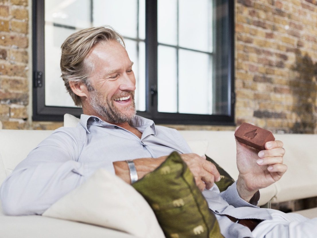 Man with miniature brick sitting on sofa in front of brick wall