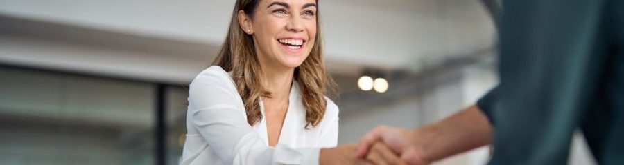 Happy mid aged business woman manager handshaking greeting client in office. Smiling female executive making successful deal with partner shaking hand at work standing at meeting table.